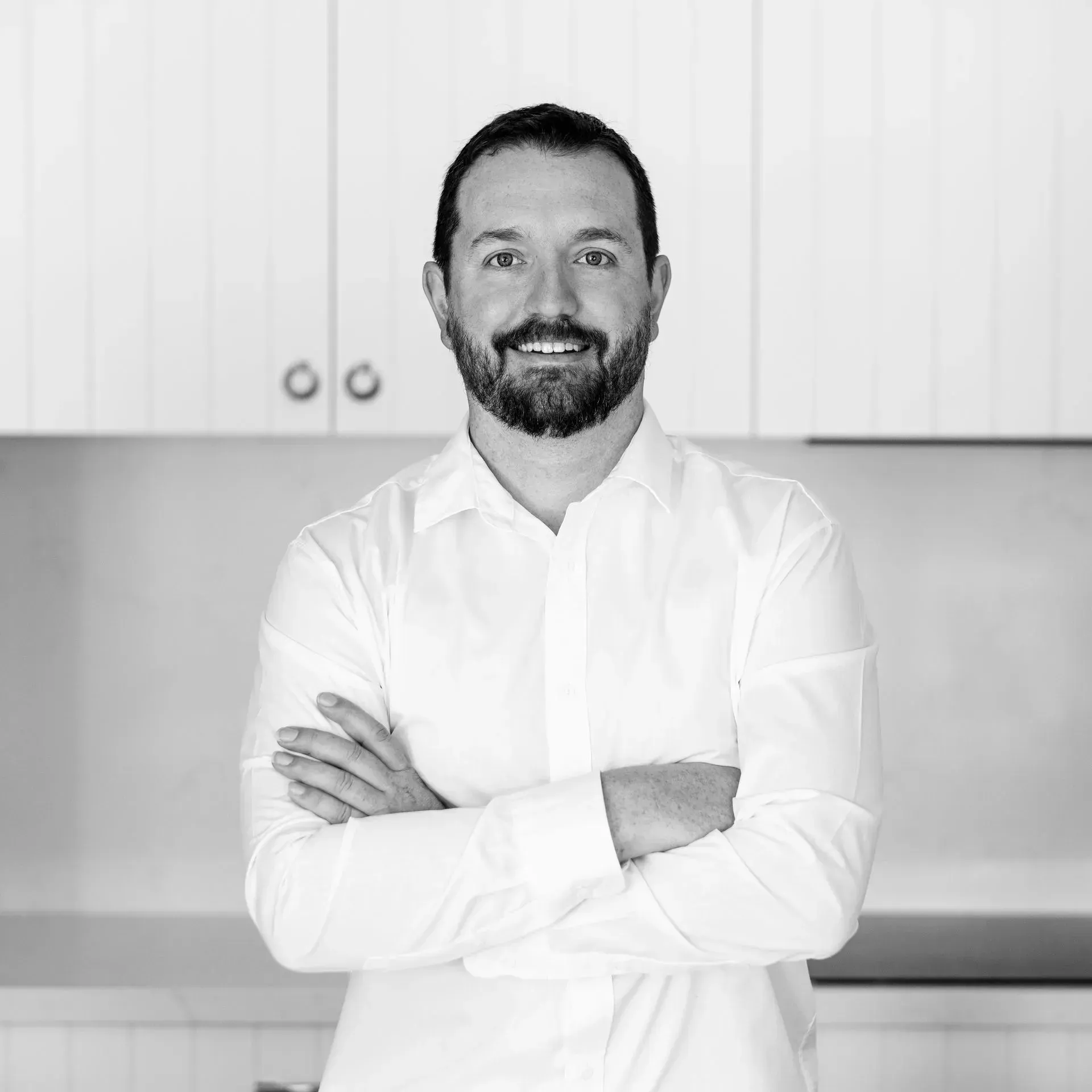 A man with a beard is standing in a kitchen with his arms crossed.