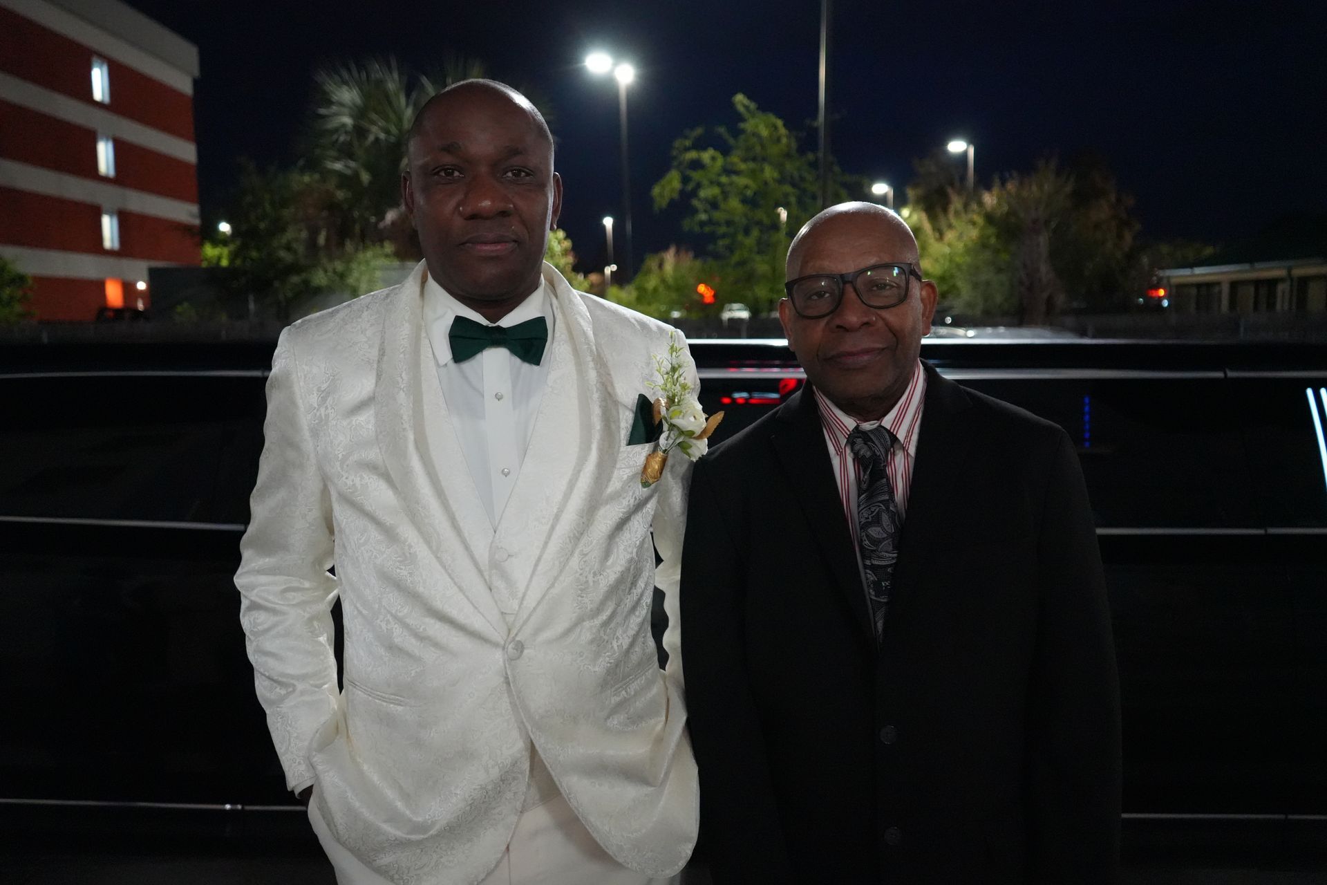Two men in suits are standing next to each other in front of a limousine.