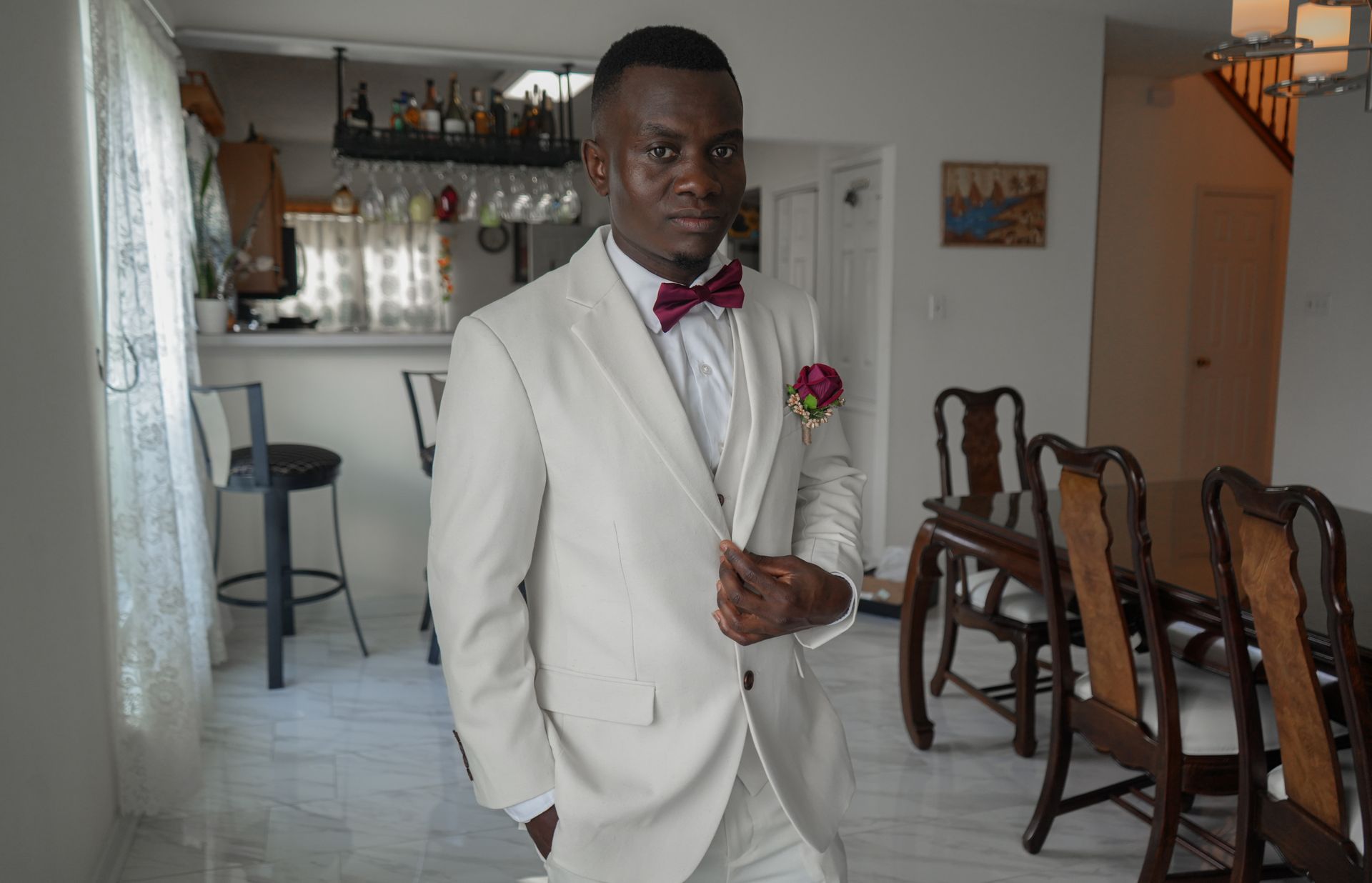 A man in a white suit and red bow tie is standing in a living room.