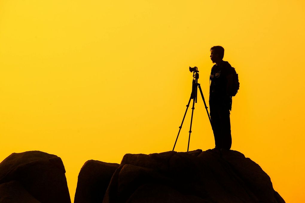 A man is standing on top of a rock taking a picture with a camera on a tripod.