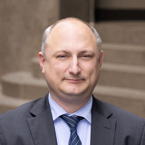 Man in suit and tie, smiling, outdoors in front of stone steps.