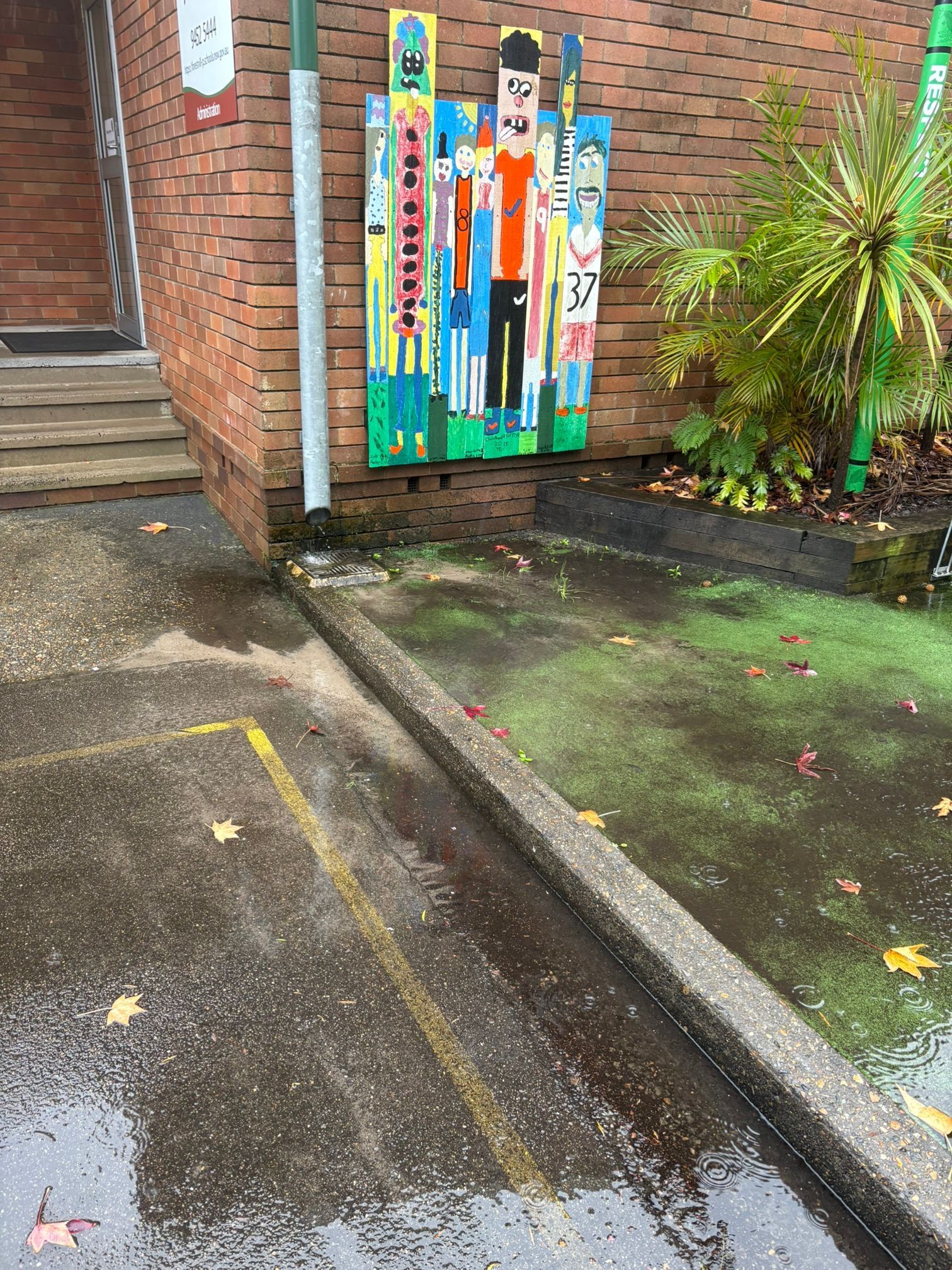 A colorful, abstract mural on a brick wall next to a puddle, a green plant, and a paved walkway on a rainy day.