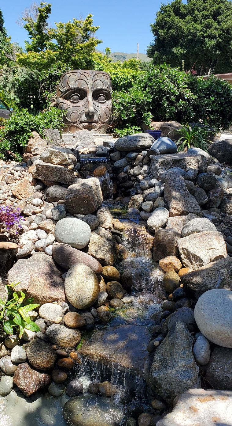 A waterfall in a garden with a statue of a face in the background.