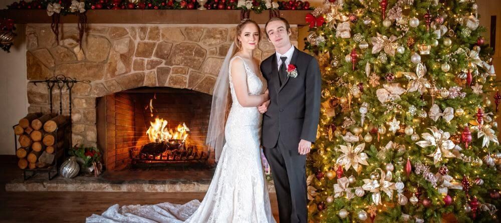 A bride and groom are standing in front of a christmas tree in front of a fireplace.