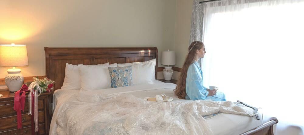 A woman is sitting on a bed in a bedroom looking out a window.