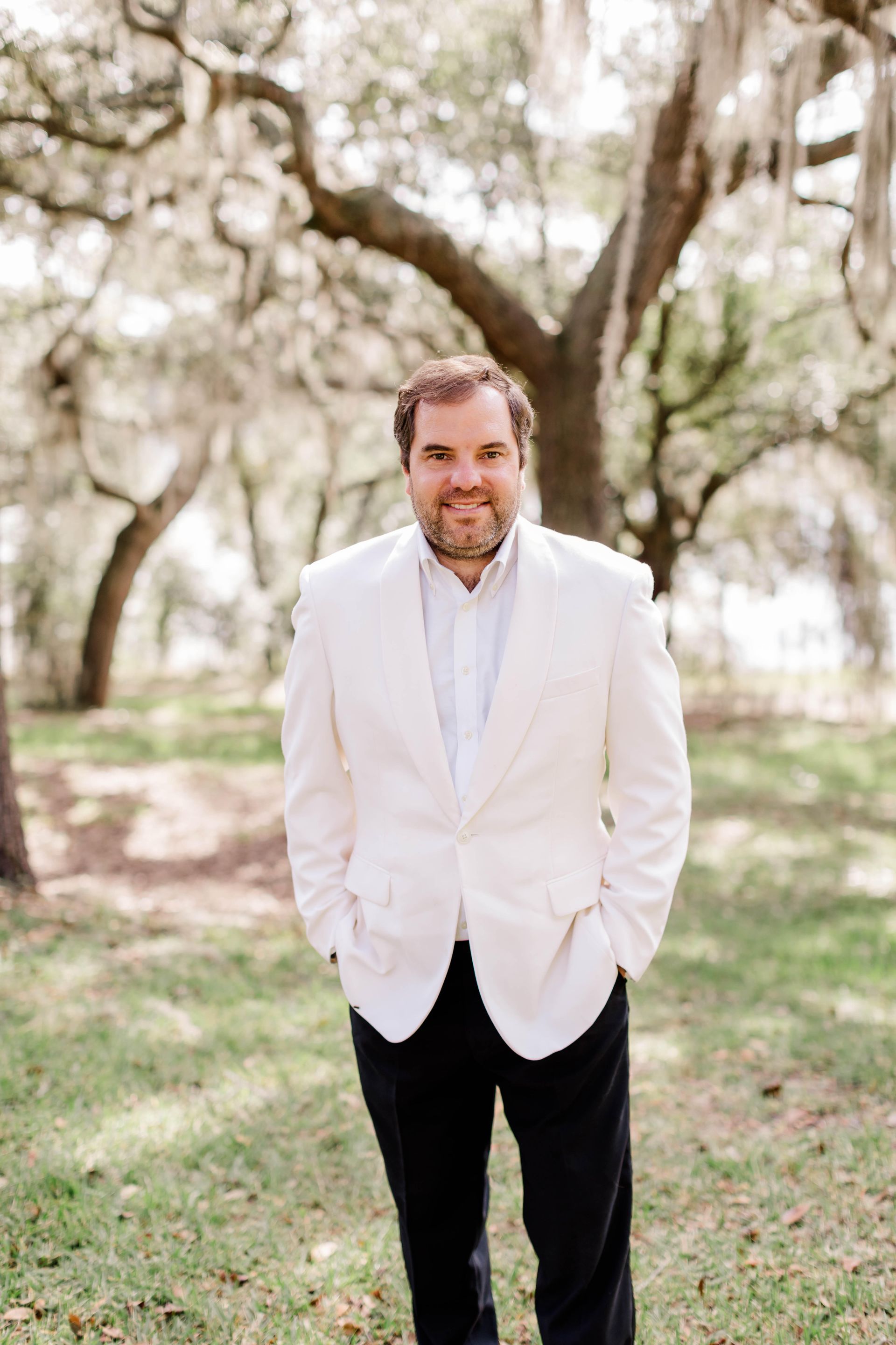 A man in a white jacket and black pants is standing in a park with his hands in his pockets.