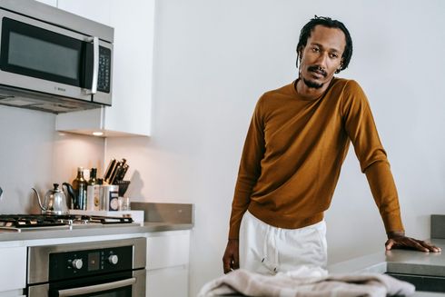 A man is standing in a kitchen leaning on a counter ,ad about the oven.