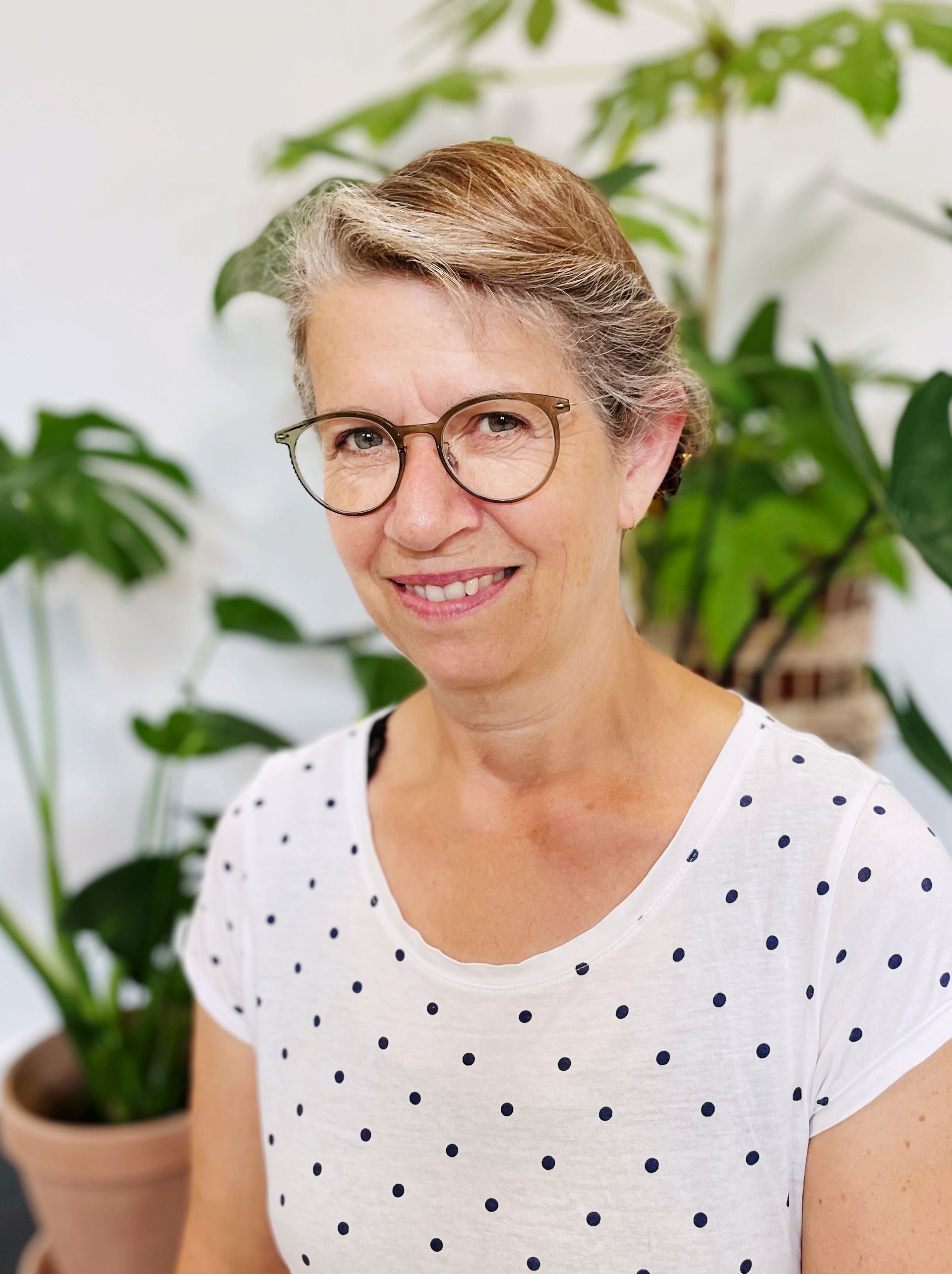 Underviser Sussie iført briller og en hvid bluse med sorte prikker smiler foran potteplanter.
