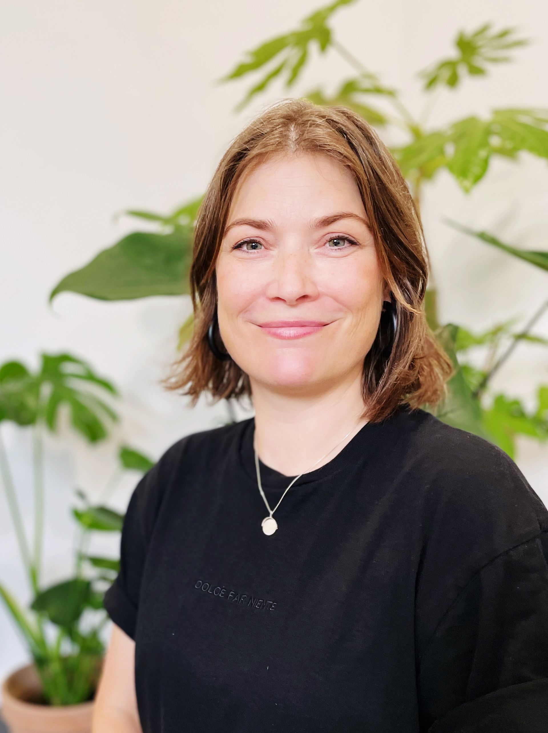 Underviser Mette har paigehår og er iført en sort bluse og en halskæde smiler foran en potteplante.