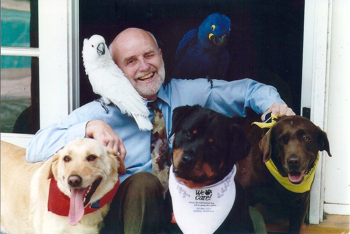 Man with dogs and parrots; he smiles with a cockatoo on his shoulder.