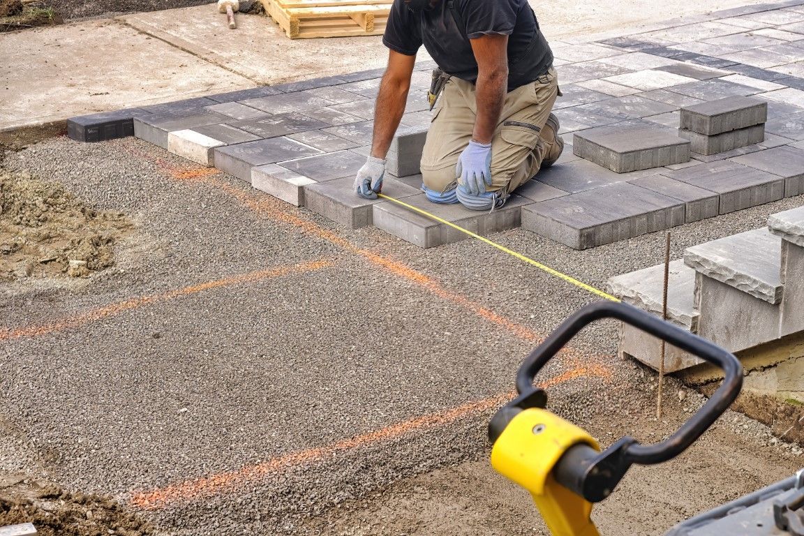 Man laying paving stones, kneeling, measuring with a tape measure. Construction site with gravel and bricks.
