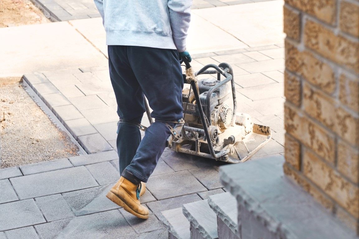 Person operating a plate compactor on brick pavement near a building.