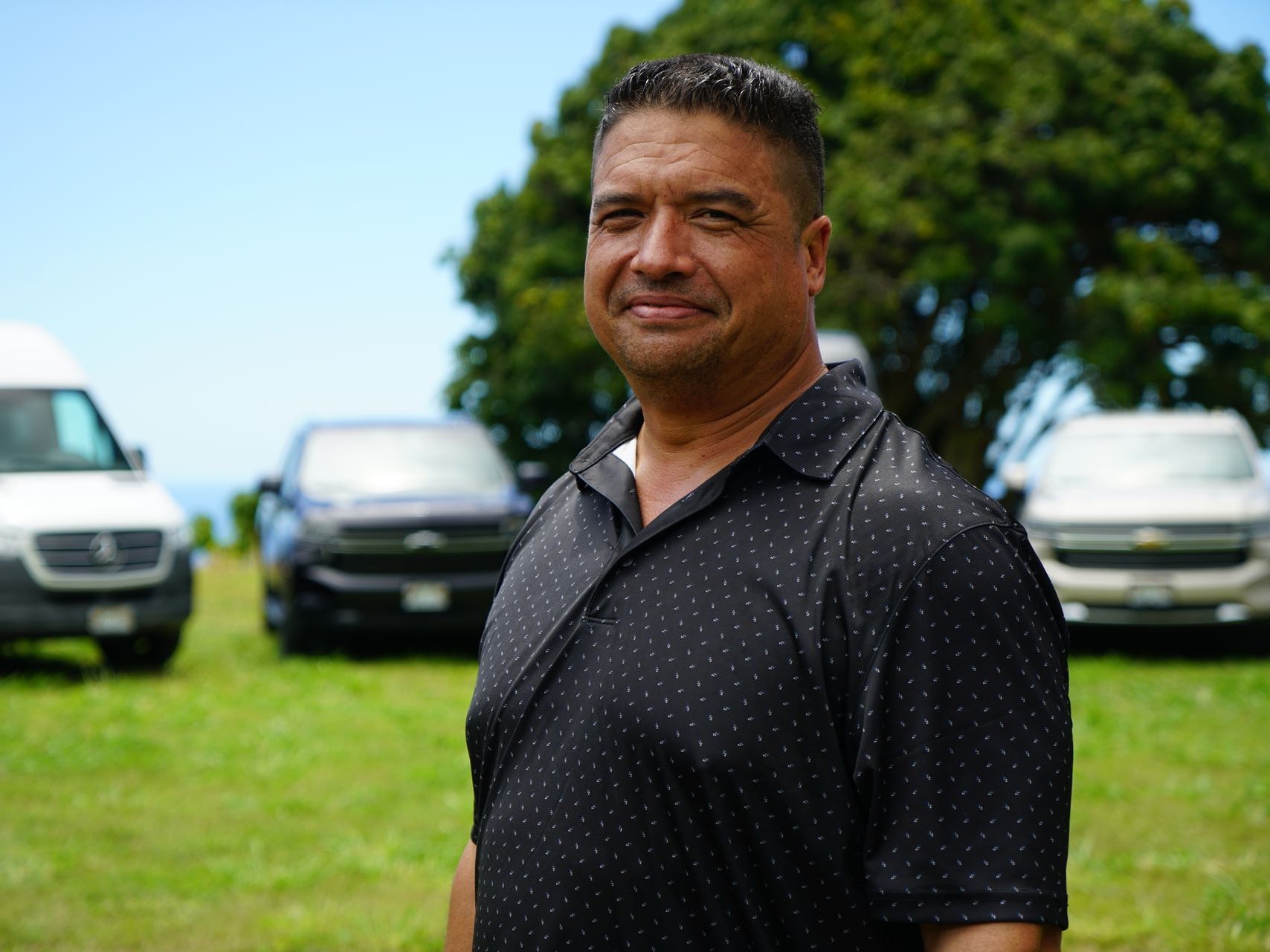 A man in a black shirt is standing in a field with cars in the background.