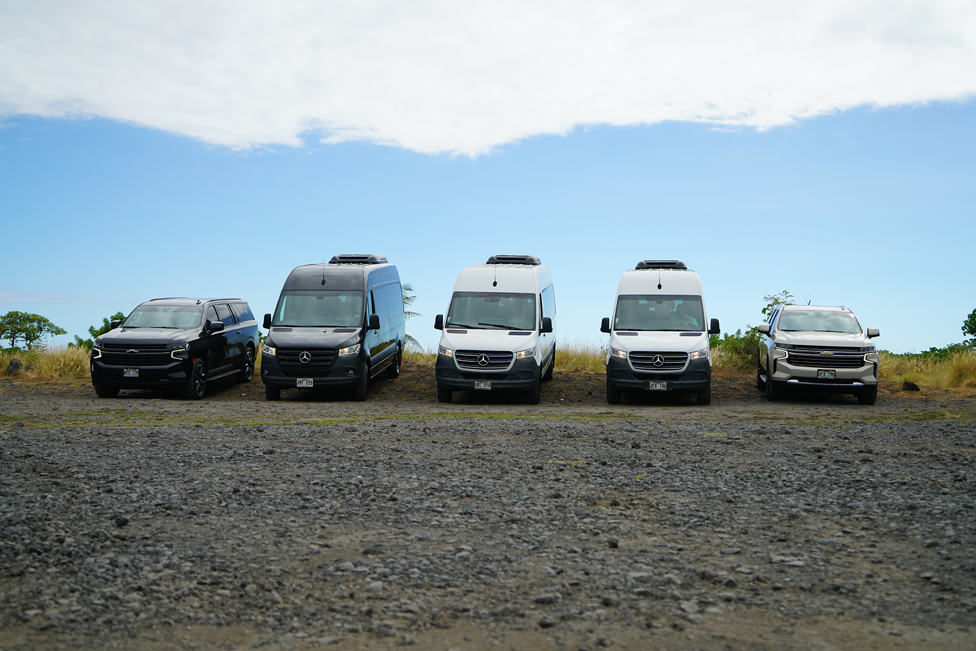 A row of vans and trucks are parked in a gravel lot.