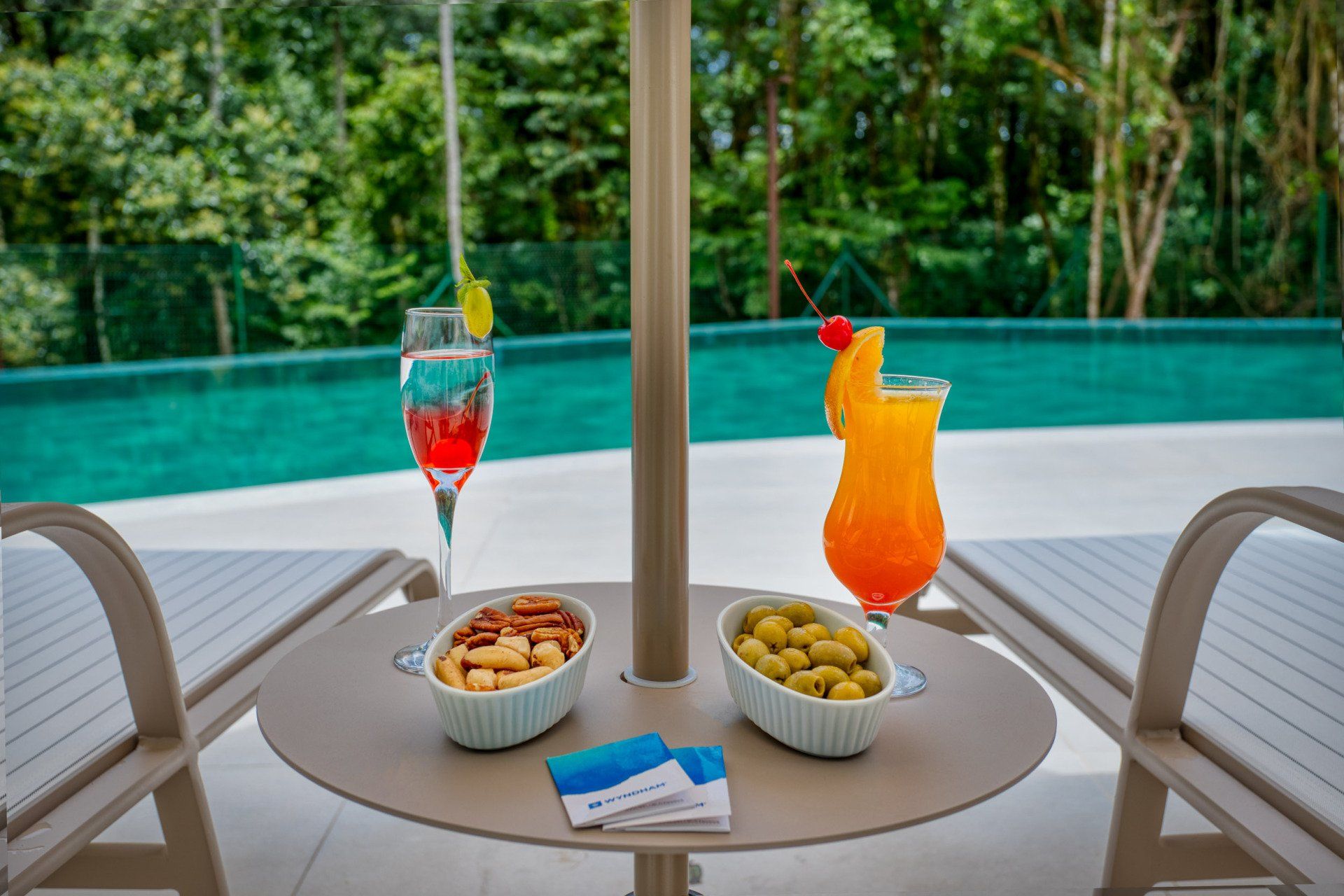 Two lounge chairs, drinks, and snacks by a pool under an umbrella.