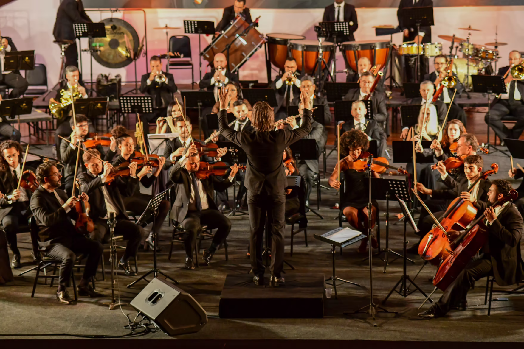 An orchestra performs outdoors at night, conductor with arms raised, lit stage, musicians playing instruments.
