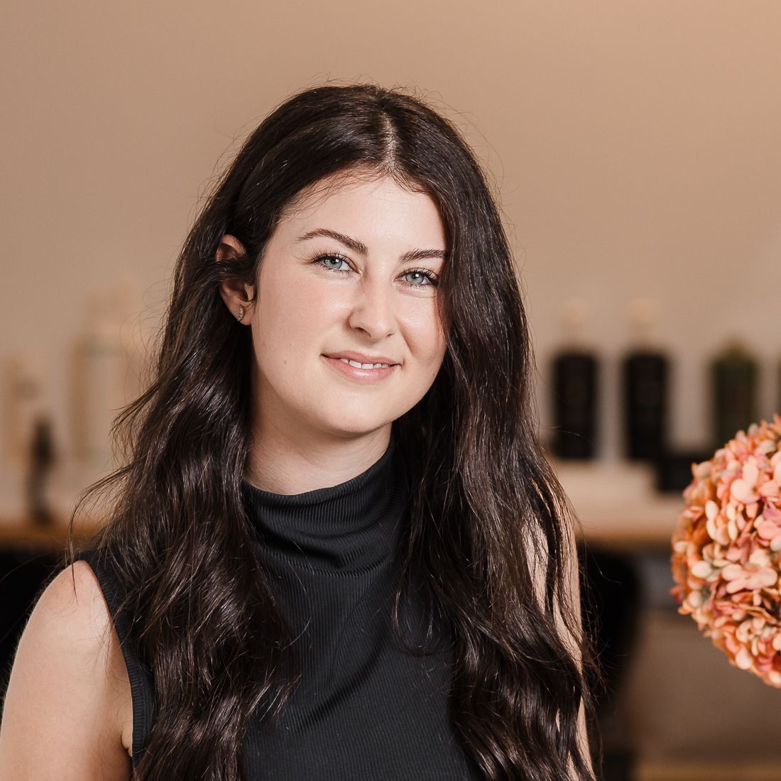 A woman in a black dress is standing in a salon next to a vase of flowers.