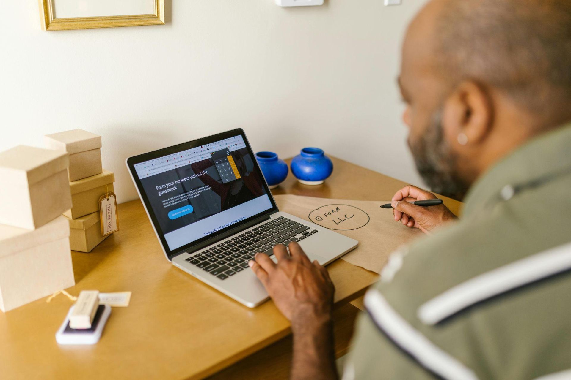 A person working on a laptop at a desk with stacked boxes, a notepad, and blue vases.