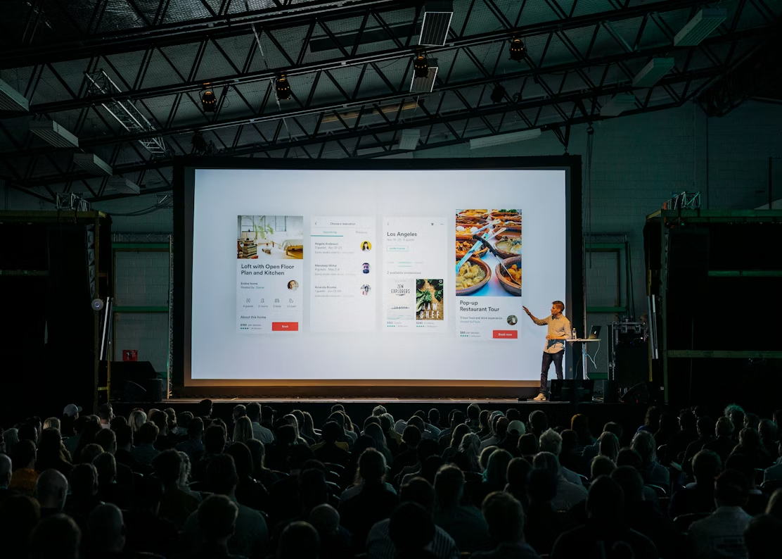 Man presenting app screens on a large screen at a conference to a seated audience.