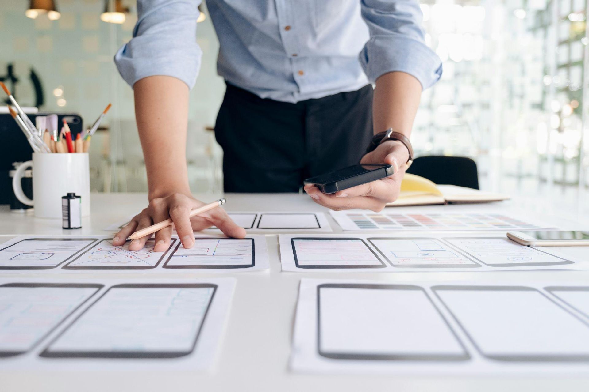 Person designing website foundation (wireframe), using pencil and phone, at desk.