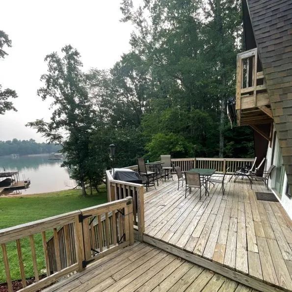 Wooden deck with outdoor furniture overlooking a lake. Trees and a house visible in the background.