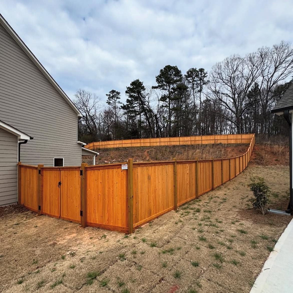 Wooden fenced backyard with a raised section beside a house and leafless trees beyond