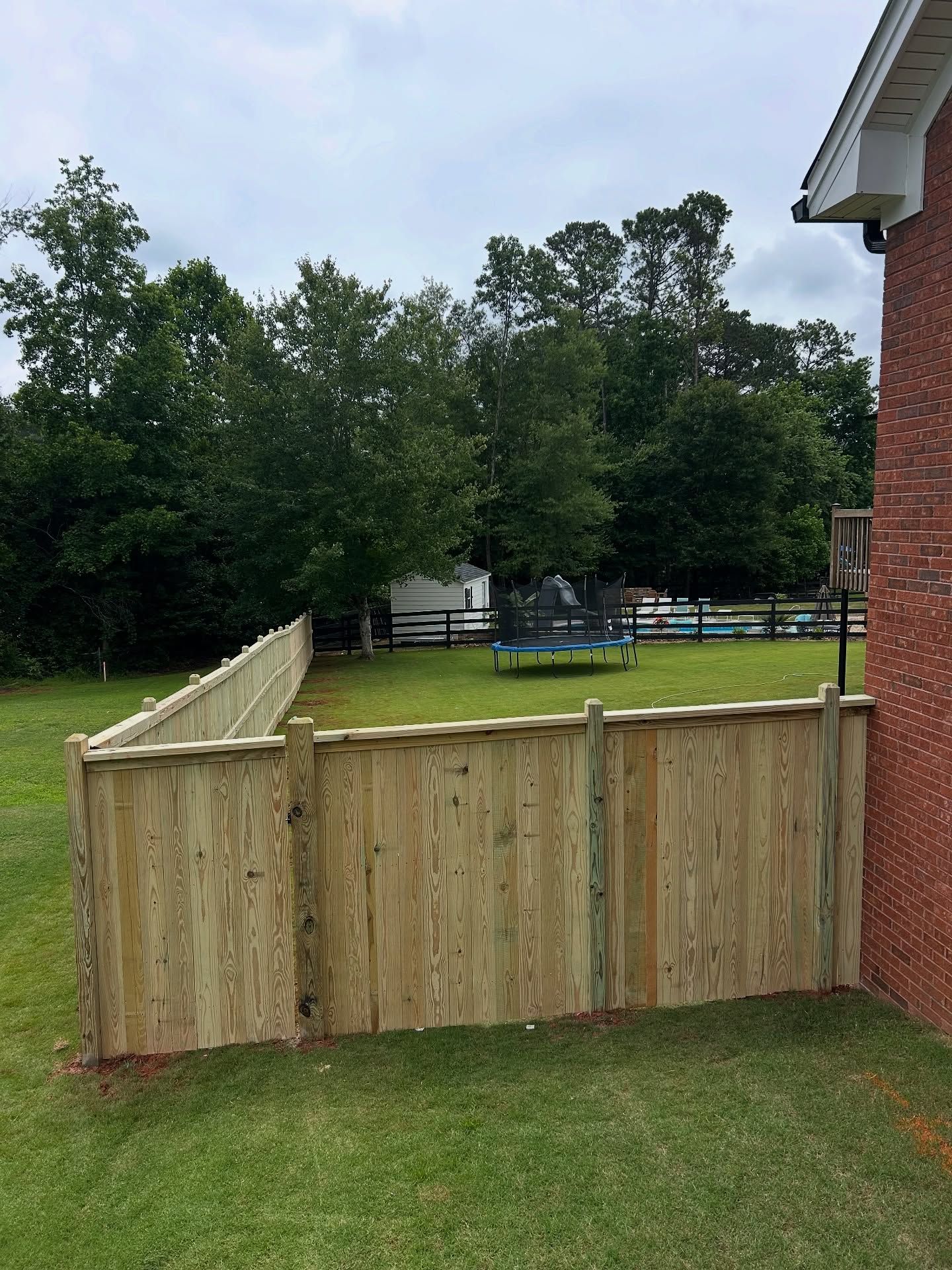 Wooden fence in a backyard with a trampoline, trees, and a brick house. Green grass and overcast sky.