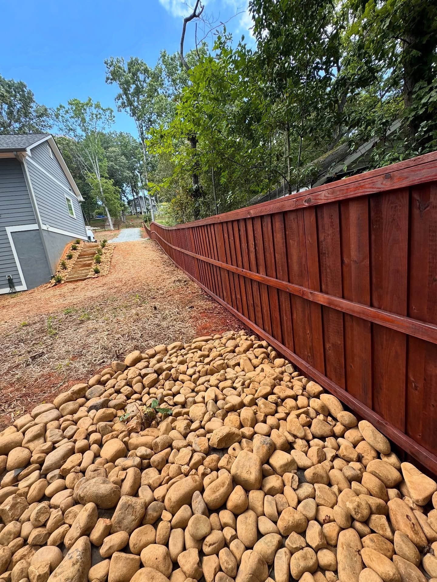 Red wooden fence beside a rocky drainage path with a gravel lane and trees along a house