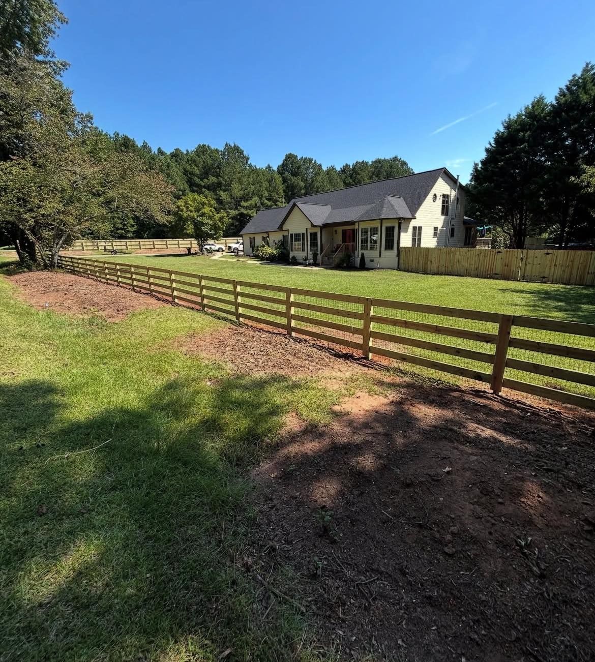 Wooden fence in front of a yellow house with a dark roof on a sunny day.