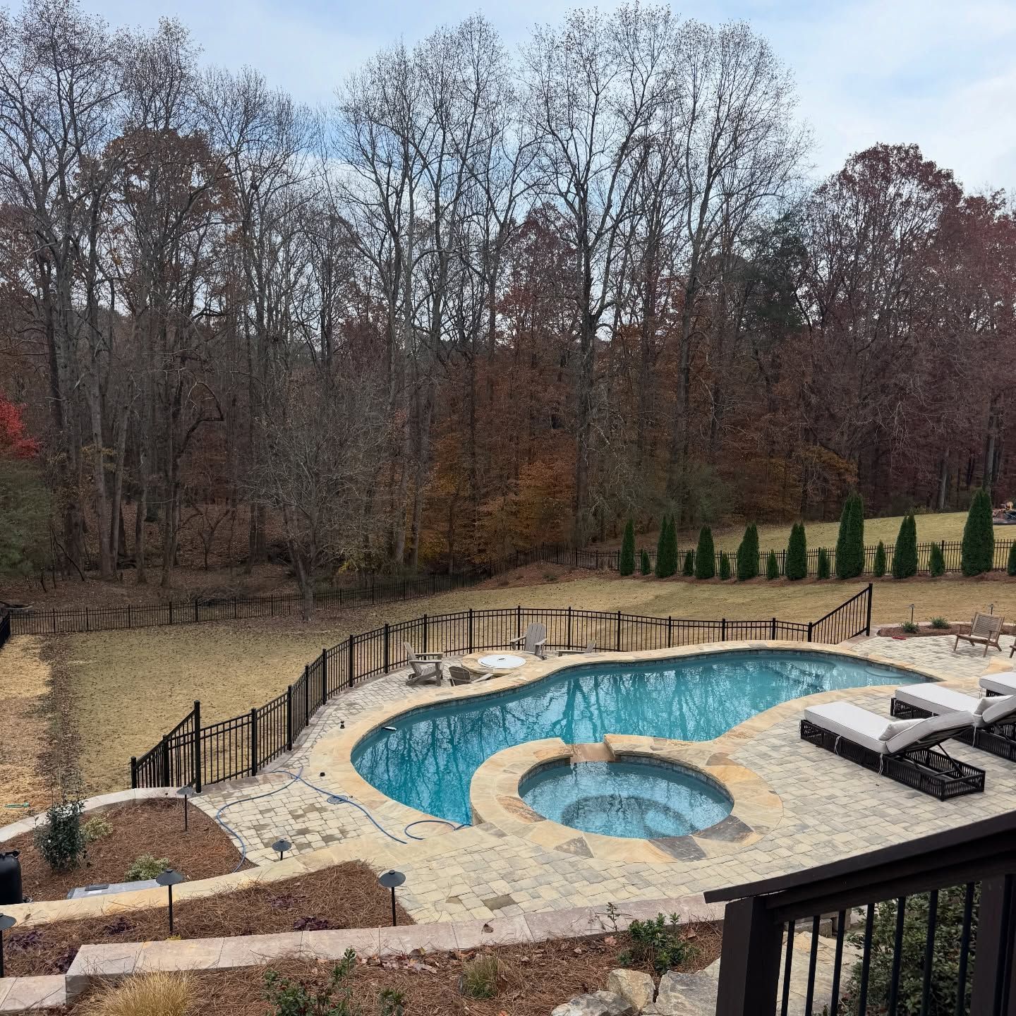 Backyard pool with stone deck, hot tub, black railing, and wooded autumn backdrop