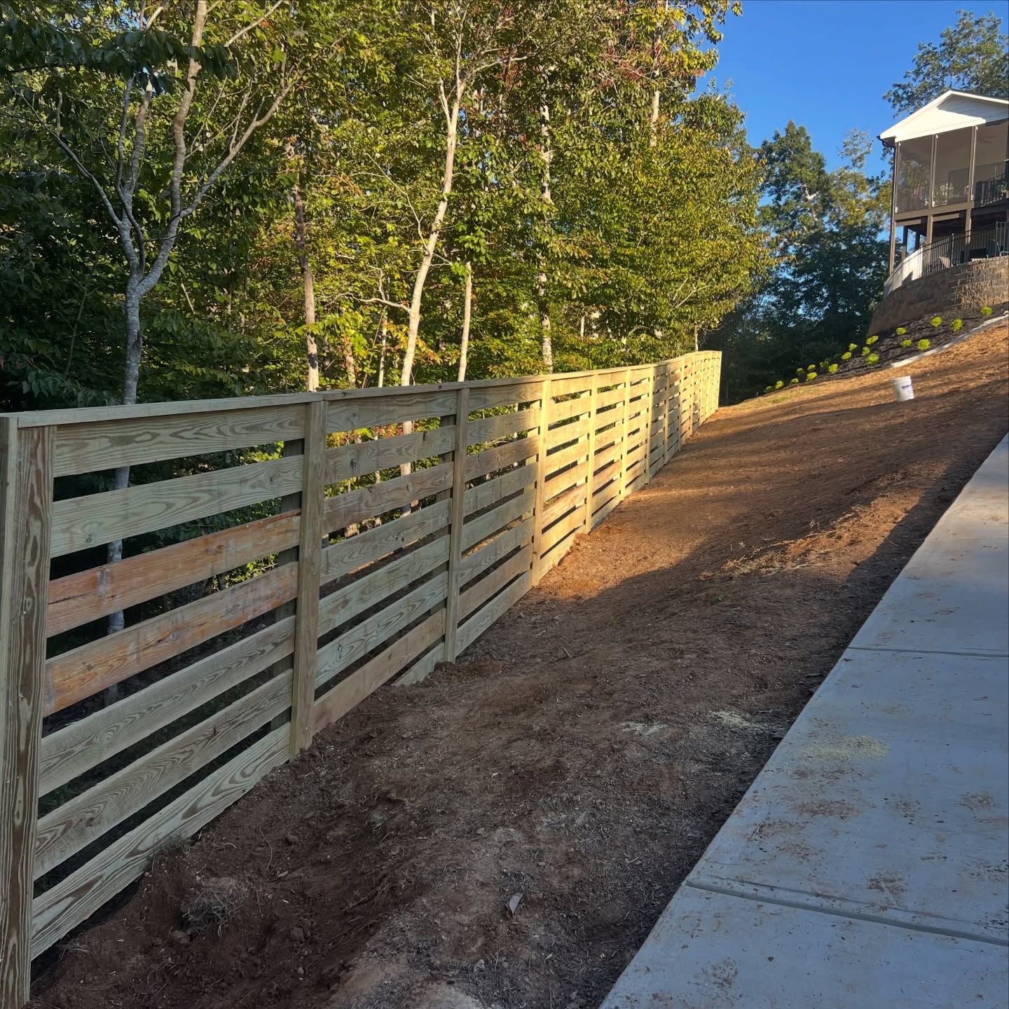 Wooden fence along a dirt hillside beside a concrete sidewalk in late afternoon sunlight