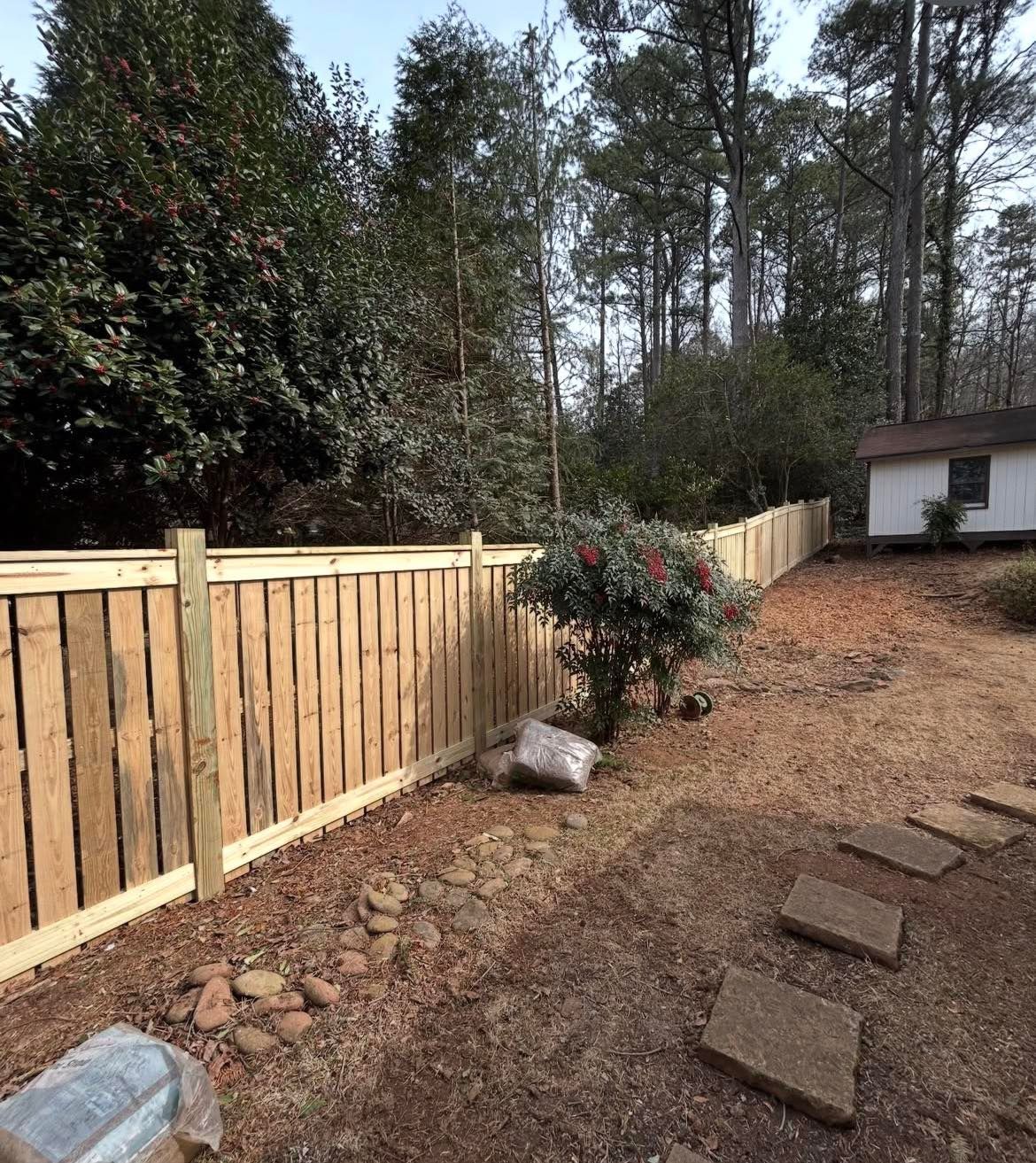 Wooden fence beside a gravel yard with stepping stones, trees, and a small white shed in the background