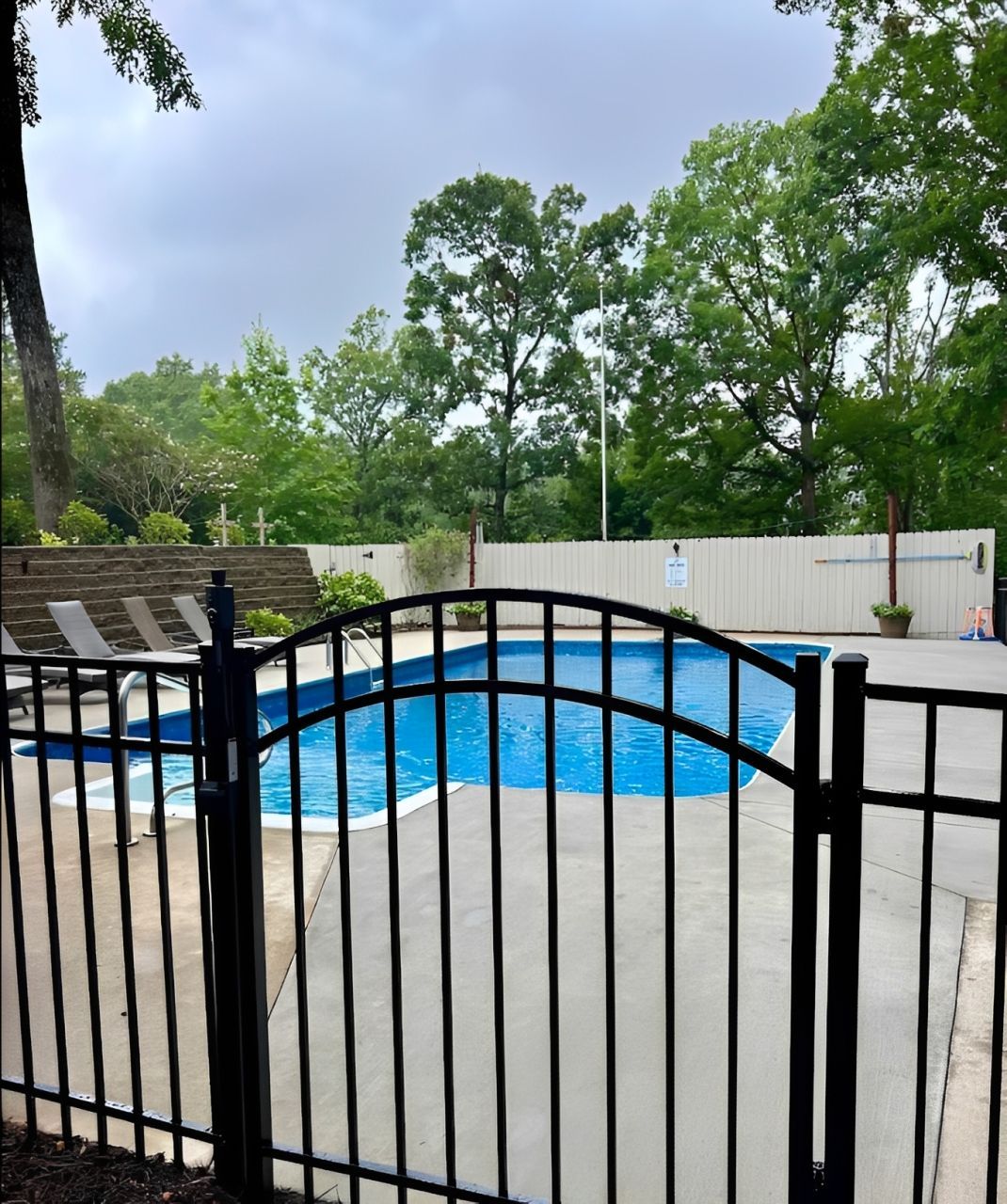 Backyard swimming pool behind a black metal fence, with trees and a white privacy fence.