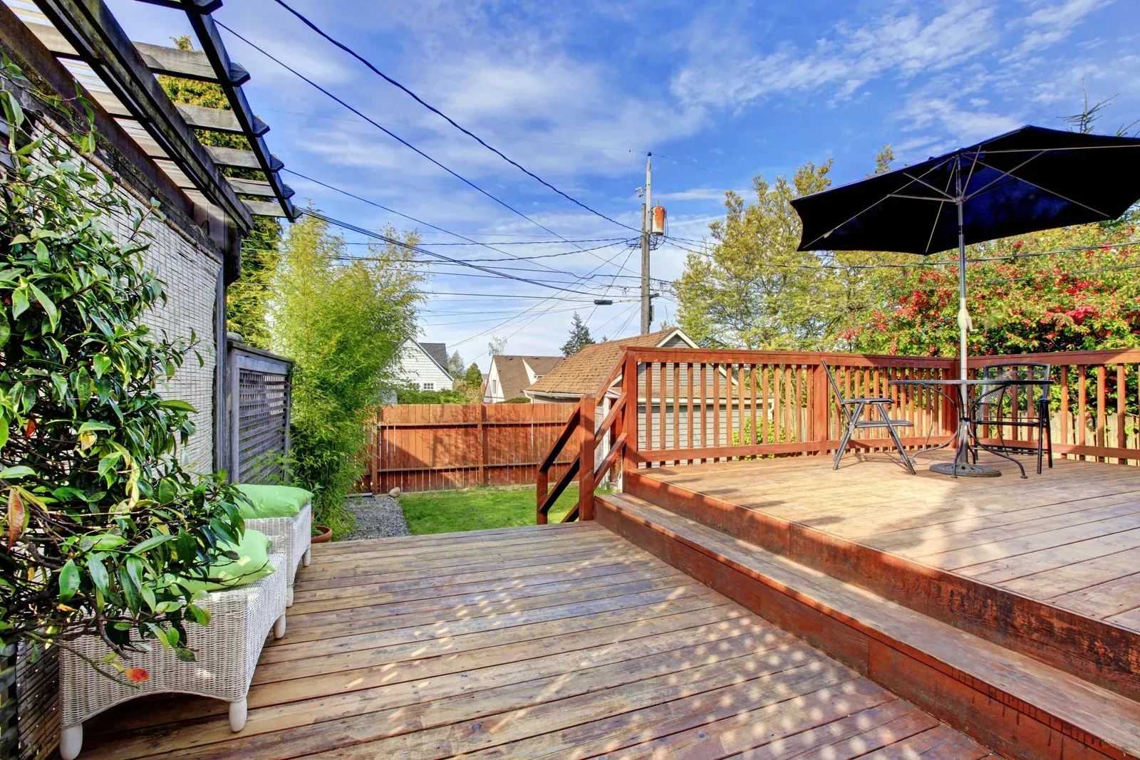 Wooden deck with a black umbrella, seating, and a fenced yard under a blue sky.
