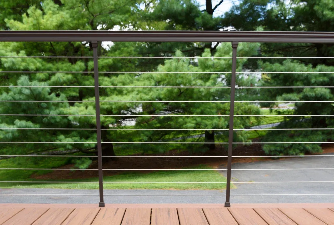 Brown deck railing with horizontal cables against a backdrop of trees, lawn, and road.