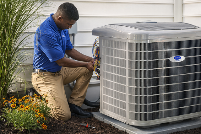 A technician in a blue uniform kneels outside, performing maintenance on a residential air conditioning unit.