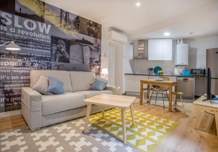 Luminoso salón moderno con sofá gris, alfombra estampada, mesa de centro y zona de cocina abierta.