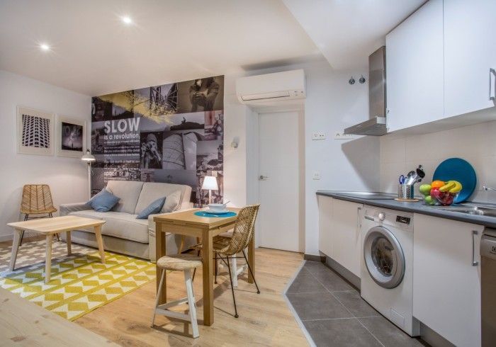 Apartamento tipo estudio compacto con sofá cama, alfombra amarilla, cocina americana y lavadora bajo un techo inclinado.