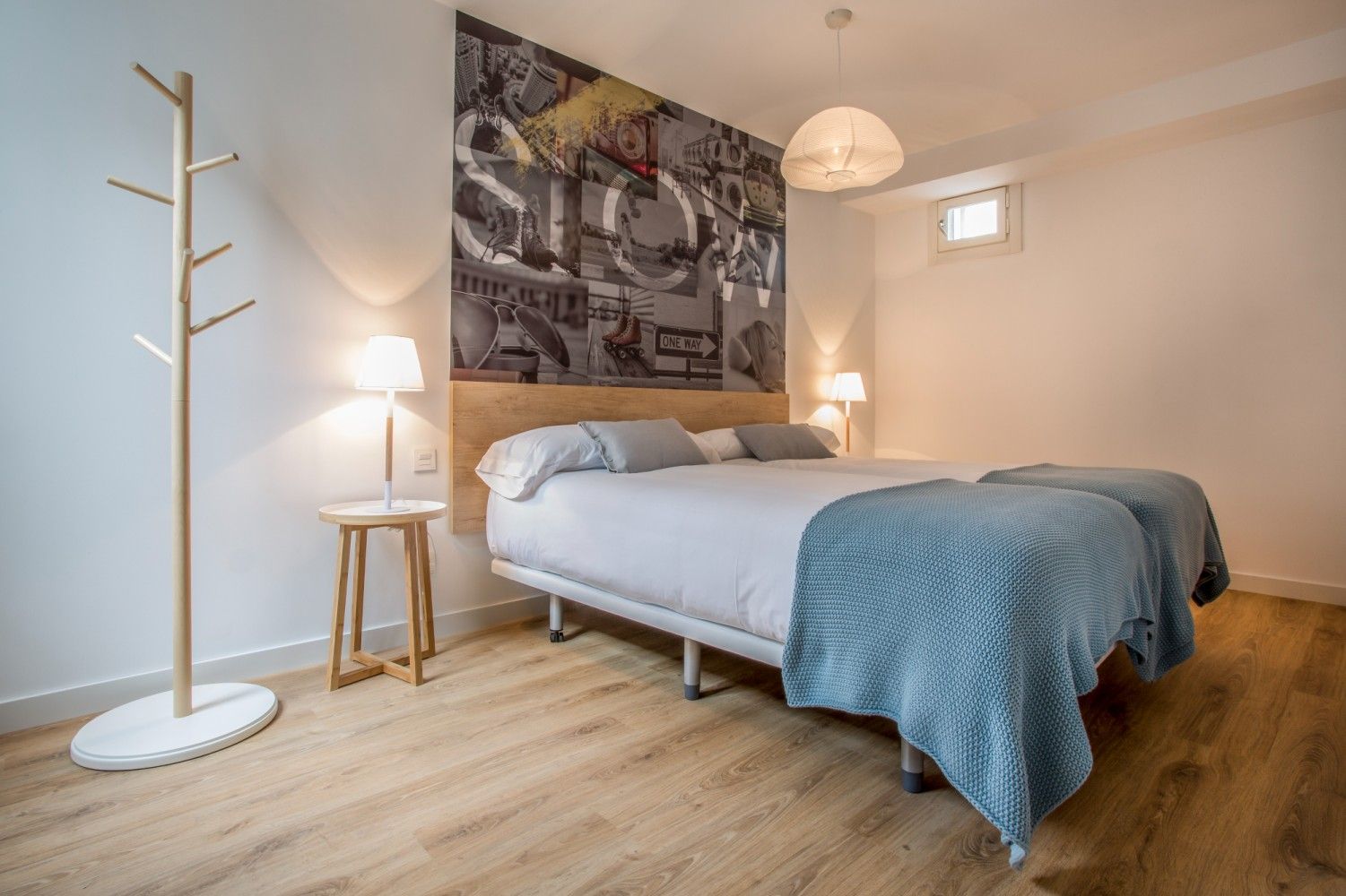 Habitación luminosa con cama, manta azul, suelo de madera, lámparas y cuadros en la pared.