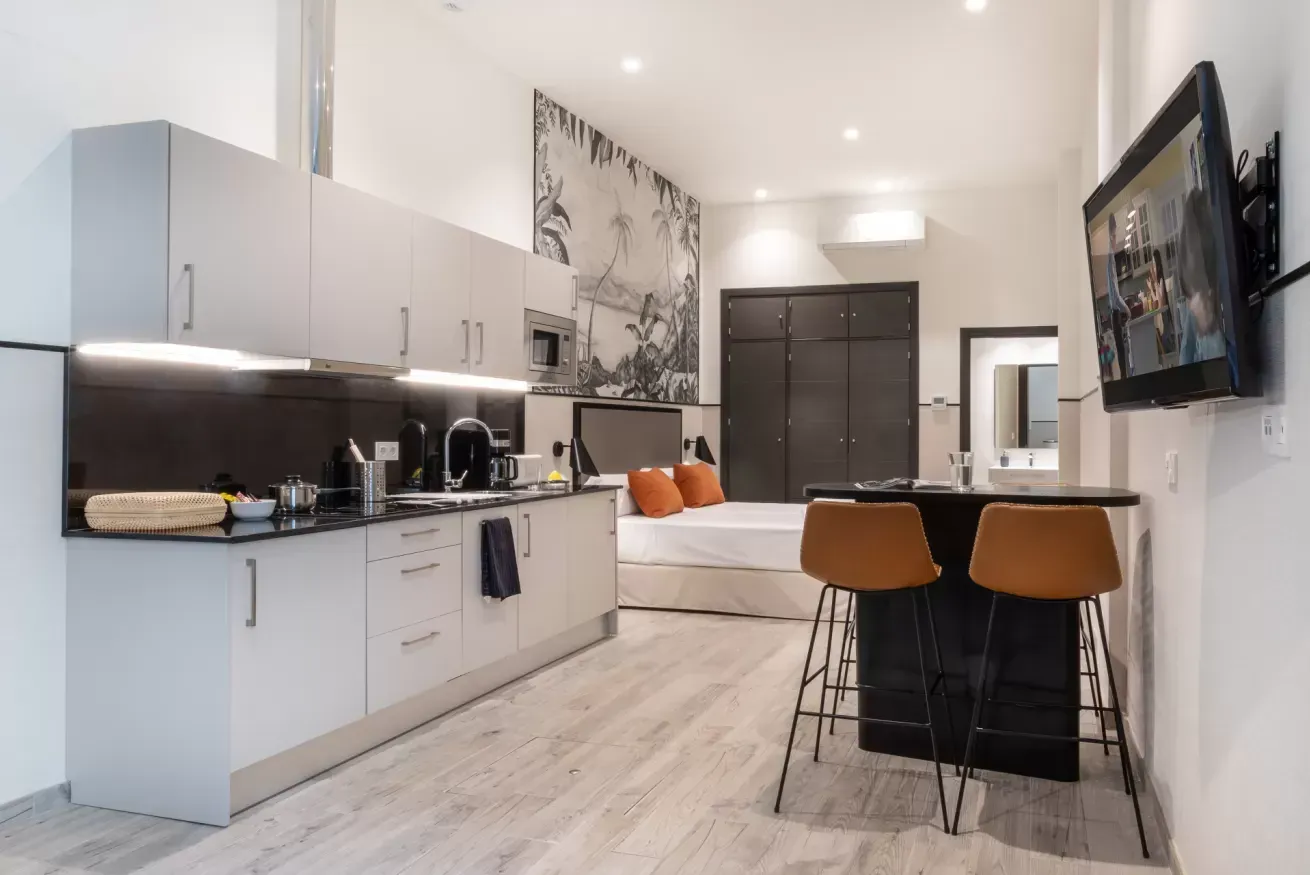 Cocina y comedor modernos de planta abierta con armarios blancos, detalles en negro y un televisor montado en la pared.