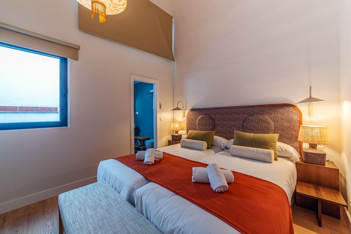 Dormitorio moderno con una cama grande, manta naranja, lámparas de noche y una puerta azul con vista al mar.