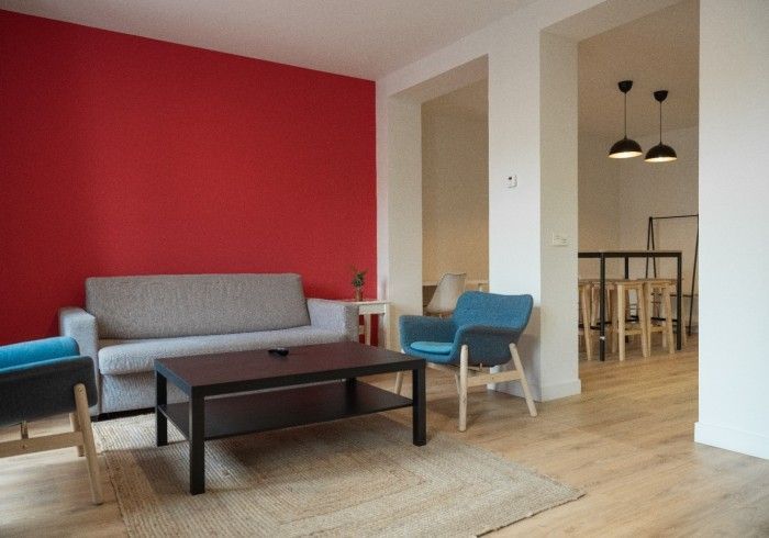 Sala de estar moderna con pared decorativa roja, sofá gris, sillón azul, mesa de centro y zona de comedor al fondo.