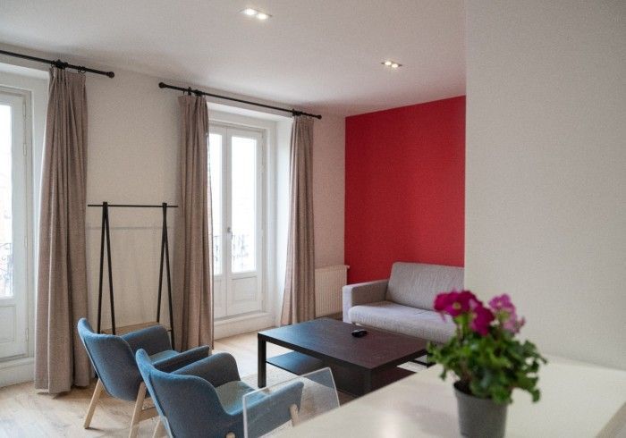 Sala de estar luminosa con sillas azules, mesa de centro negra, pared decorativa roja y ventanas altas con cortinas.
