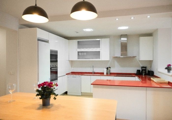 Cocina moderna y luminosa con armarios blancos, encimeras rojas y una mesa de comedor bajo lámparas colgantes.
