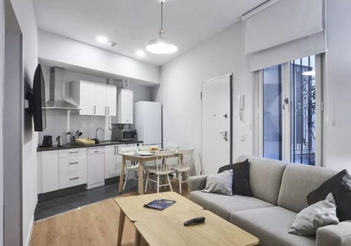 Apartamento tipo estudio moderno con sofá gris, mesa de madera, cocina compacta y puerta de balcón junto a una ventana.