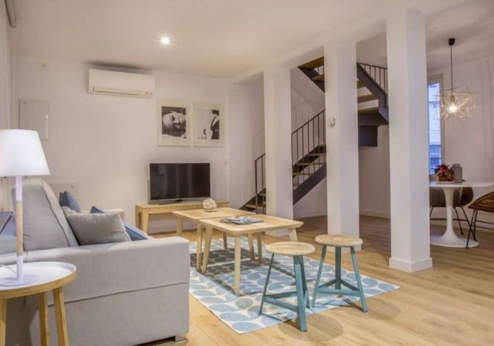 Sala de estar luminosa con sofá gris, mesas de madera, alfombra estampada, escaleras y cuadros en la pared.