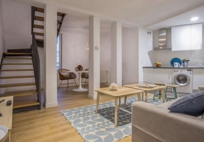 Apartamento moderno de planta abierta con escalera, cocina americana, mesa de comedor y sofá en tonos neutros.