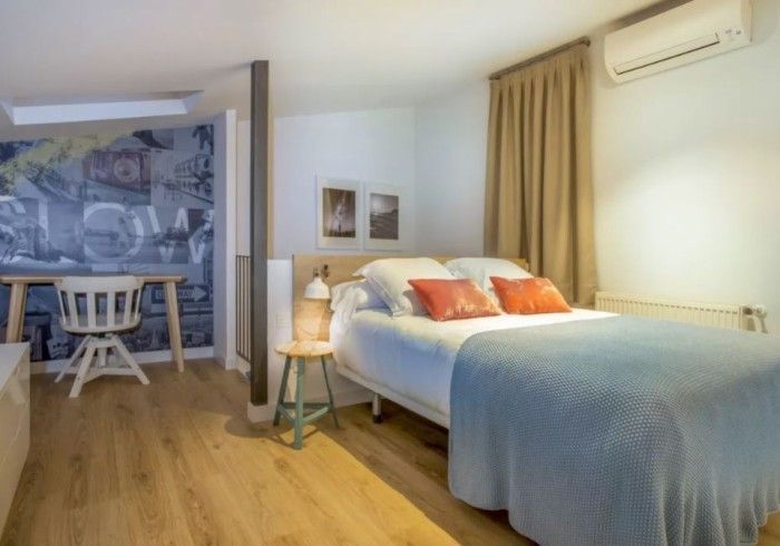 Habitación luminosa con cama doble, manta azul y una pequeña zona de comedor cerca de una gran ventana.