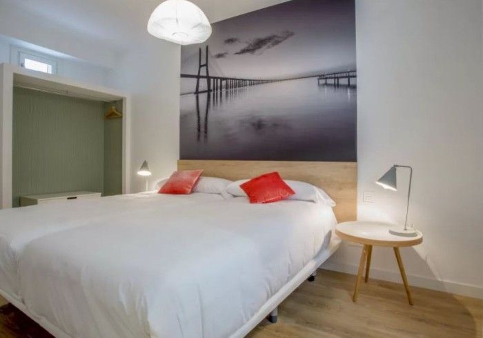 Habitación luminosa con cama blanca, almohadas rojas, lámpara de noche y un gran cuadro en blanco y negro.
