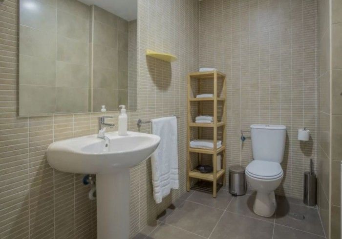 Baño pequeño con lavabo, espejo, toallero, estantes de madera, inodoro y paredes de azulejos beige.